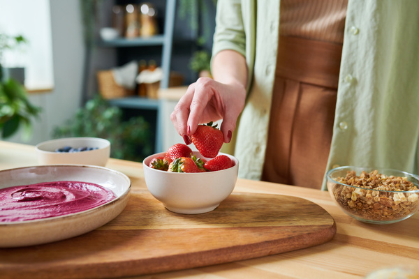 The smoothie maker takes a strawberry from a white bowl standing on a wooden serving board between a dish with a pink smoothie and a transparent bowl with granola