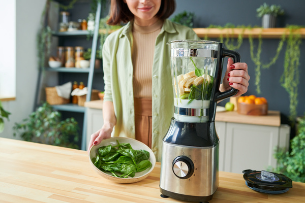 A smoothie maker with dark short hair and light clothes holds a dish with spinach leaves in one hand and holds the handle of a bender with the other in a jug of which there is a sliced banana and spinach