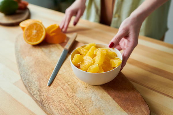 A woman is going to take a white bowl full of sliced orange from a wooden cutting board on which there is a knife  half an orange with a peel