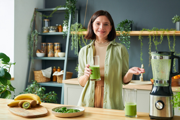 A female with a short haircut in a light outfit smiling and pointing to the blender holds a glass of smoothie in her hand standing at the kitchen table with fruit and a plate of spinach on it