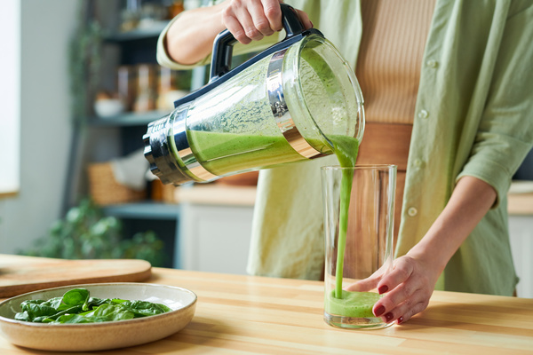 A smoothie maker in light clothes pours a green fruit and spinach drink from a jug from a stationary blender into a glass standing on the table next to the dish on which the greens are lying