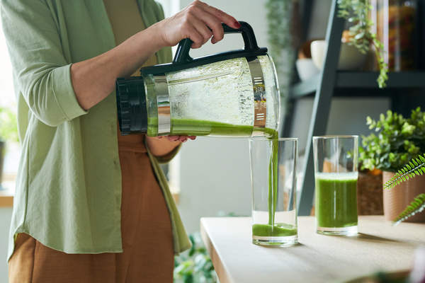 A smoothie maker in a light green shirt standing in the kitchen pours a green smoothie into a glass from a jug of a blender with a black handle