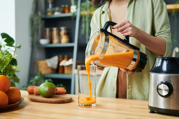A woman in light clothes pours a bright orange smoothie from a jug from a blender standing next to her into a glass