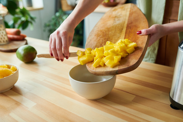 A woman who makes smoothies rakes diced mango from a board with a knife into a white bowl