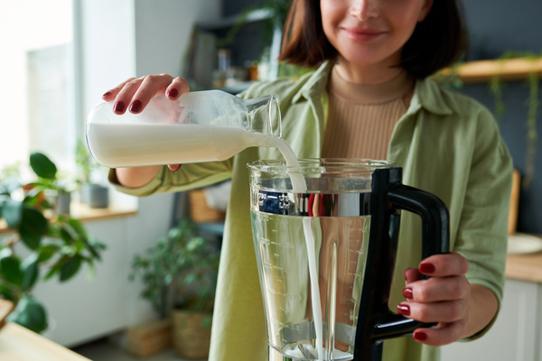 A woman making a smoothie pours milk from a glass bottle into a blender jug while standing in the kitchen with potted plants