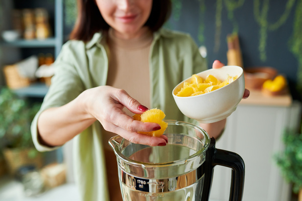 A smoothie maker in a light outfit with short hair adds orange slices from a white bowl to a jug of a stationary blender