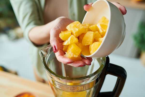 A woman who makes smoothies dressed in a light outfit pours a sliced orange from a white bowl into a jug of a stationary blender