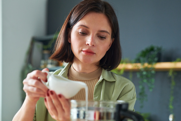 A woman with short dark hair cooking a smoothie carefully pours milk from a white gravy boat into a blender jug