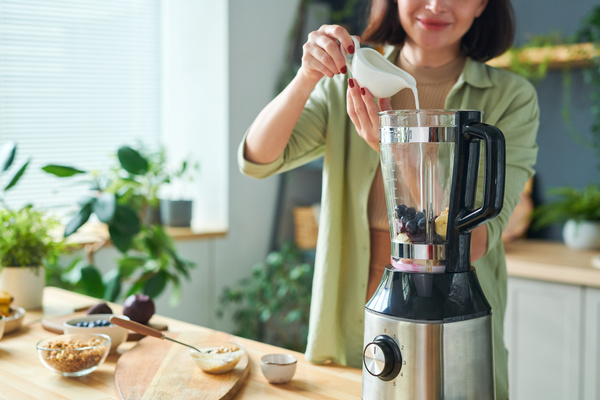 A female smoothie maker in light clothes with short dark hair pours milk from a white gravy boat into a jug of a blender containing sliced banana and blueberries