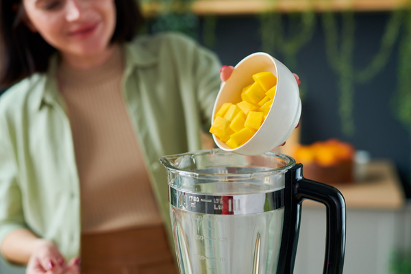 A female smoothie maker pours sliced mango from a white bowl into a blender jug