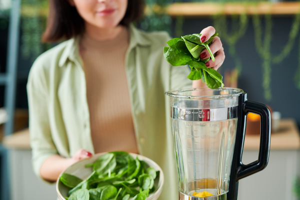 A smoothie maker with a short haircut dressed in light clothes throws a handful of greens from a white plate into a blender jug