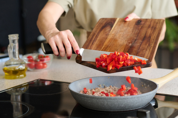 The woman holds a wooden cutting board in his hand  rakes pieces of sliced bell pepper from it with a knife into a frying pan standing on an induction stove