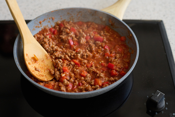 Bolognese sauce is stewing with a wooden chef's spoon left in the pan in a grey frying pan standing on a black hob