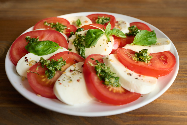 Close-up of caprese salad garnished with basil leaves and seasoned with pesto sauce served on a white flat plate