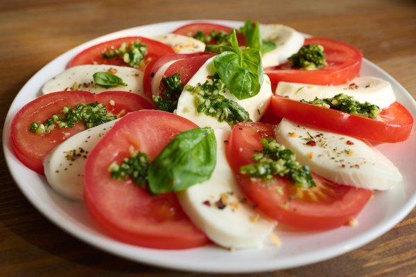 Caprese salad garnished with herbs and seasoned with pesto sauce neatly laid out a white flat dish which is on a dark wood table