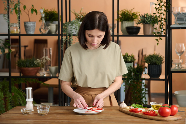 A woman cook with dark hair in beige clothes lays out a caprese salad on a plate standing at a table on which there is a board with tomatoes  garlic  basil and oil  and there are bowls with seasonings and a salt shaker