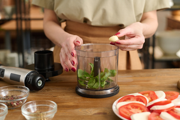 A female chef cooking basil salad sauce is going to add a clove of garlic to a blender standing on a wooden table next to a caprese salad laid out on a white dish and bowls with seasonings