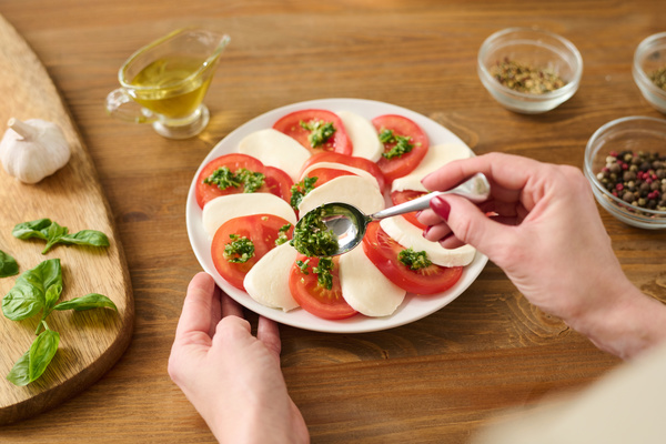 The cook puts green sauce on tomatoes in a caprese salad laid out on a plate that is on the table next to the gravy boat with butter and a cutting board with basil leaves and a garlic clove