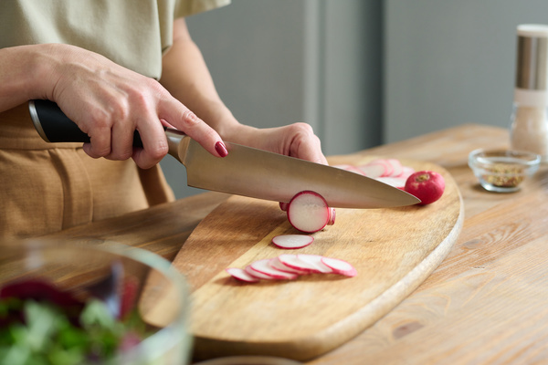A cook with red painted nails standing at a wooden table chops radish on a cutting board with a large knife
