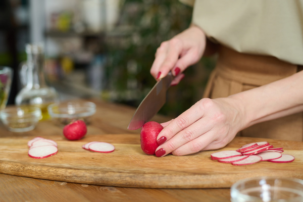 A woman cook with red painted nails stands and is about to chop a radish with a chef's knife on a wooden board with slices of radish