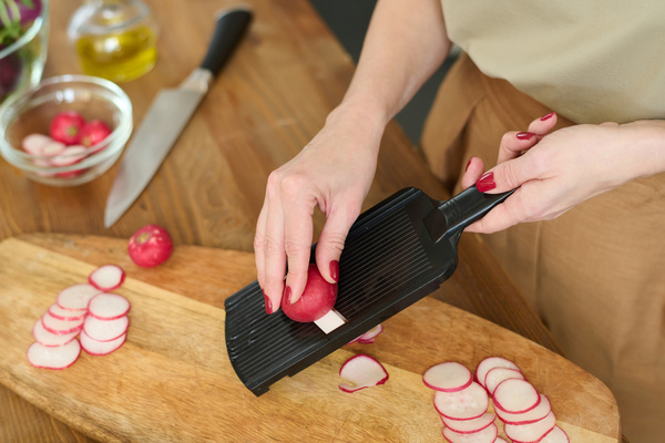 The cook standing at the table on a wooden cutting board on which there are slices of radishes is chopping radish on a black vegetable slicer