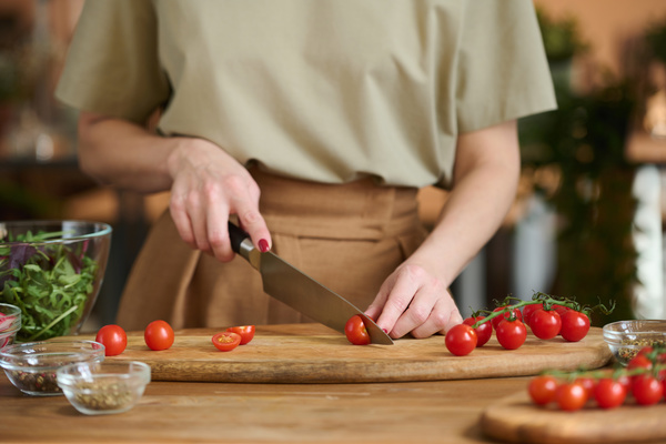 A chef in beige clothes cuts cherry tomatoes with a chef's knife on a wooden chopping board next to which there are seasonings in sauce bowls and greens in a salad bowl
