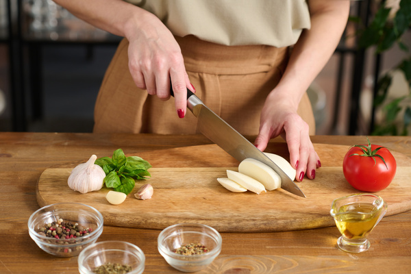 A chef in a beige outfit with red painted nails cuts mozzarella cheese with a large knife on a wooden board on which there is a tomato garlic and basil and next to it there is a gravy boat with oil and bowls with seasonings