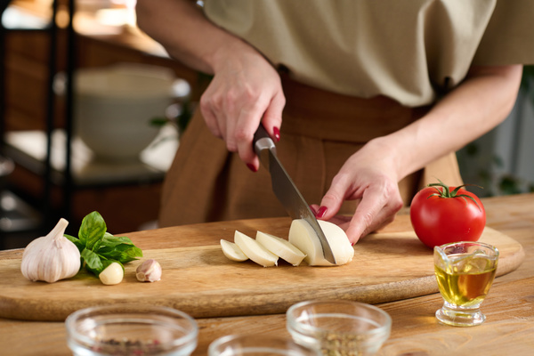 A female chef in light clothes with a red manicure  cutting mozzarella into thick slices on a wooden cutting board next to a tomato  garlic  herbs and oil in a gravy boat