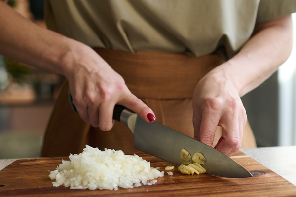 A chef in beige clothes with wine-colored nails on a board with chopped onions cuts garlic