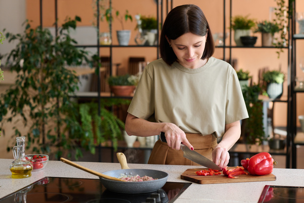 A cook with a dark square in beige clothes cuts red bell peppers on a board while minced meat with onions is fried on the cooking surface in a frying pan next to it