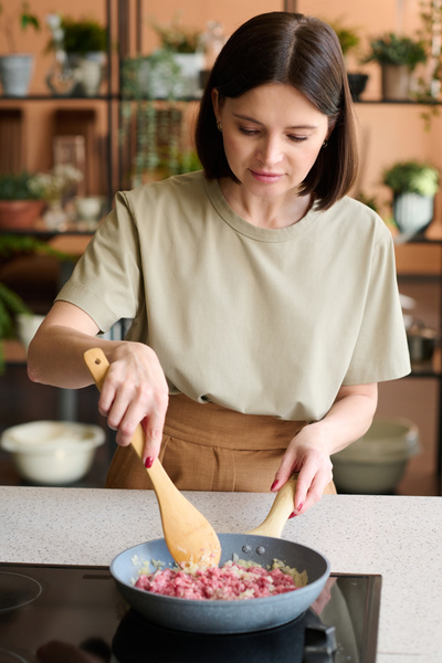 A cook with dark hair dressed in light-colored clothes is frying minced meat with chopped onion in a frying pan  stirring it with a wooden chef's spoon