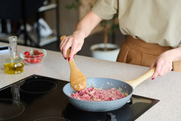 A chef with red-tinted nails using a bamboo spoon mixes minced meat fried in a gray frying pan with finely chopped onions