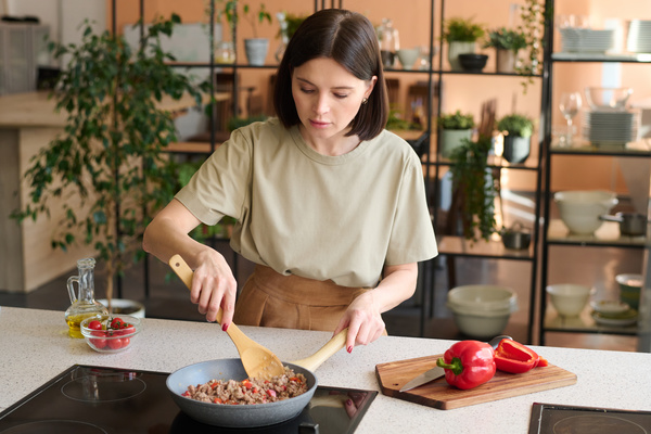 Chef with a dark square in a beige outfit with a wooden spoon is stirring minced meat with red bell pepper on the cooking surface next to the cutting board on which the pepper lies and with a bowl of cherry tomatoes and a jug of oil