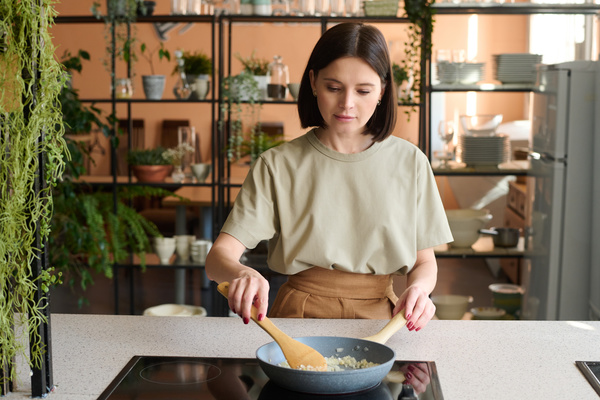A woman with a dark square dressed in beige clothes mixes finely chopped onions frying in a pan in the kitchen against the background of shelves with potted plants and dishes