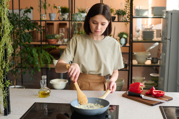 A cook with dark hair dressed in light-colored clothes is frying onions in a pan in the kitchen with shelves with potted plants and dishes on them
