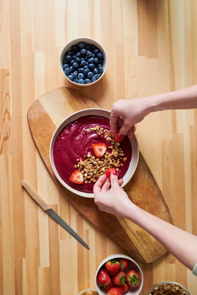 The smoothie maker adds strawberry slices to a pink smoothie strewn with granola in a deep plate standing on a wooden serving board next to blueberry and strawberry bowls