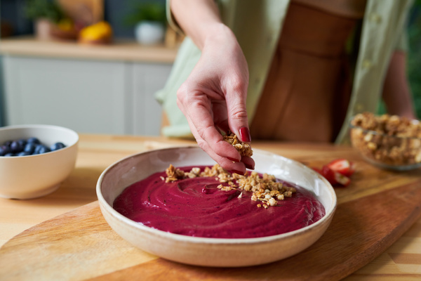 The smoothie maker sprinkles granola on a pink berry smoothie in a light bowl standing on a wooden chopping board next to a plate of blueberries