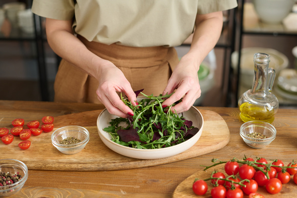 A female chef in a beige outfit lays out arugula and basil on a platter which is on a wooden chopping board next to cherry tomatoes seasonings in sauce bowls and a jug of oil on the table