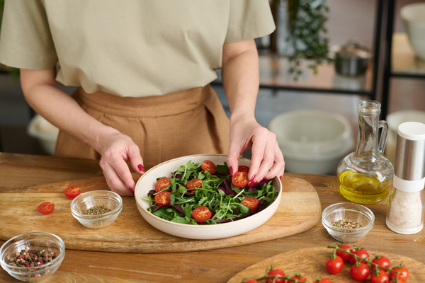 The cook  while cooking a vegetable salad  puts cherry tomatoes cut in half in a dish with salad greens standing on the table with a jug of oil and seasonings in a small bowl