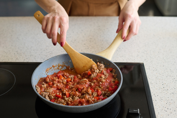 The cook is stirring the bolognese sauce with a wooden spoon  which is cooked in a gray pan