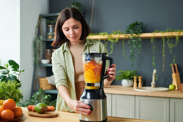 A smoothie maker with dark short hair is turning a stationary blender on in a jug of which there are sliced oranges and grated carrots to make smoothie