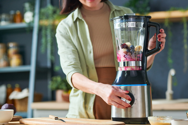 A woman in light-colored clothes is going to turn on a stationary blender in a jug of which there are fruits and berries to make a smoothie