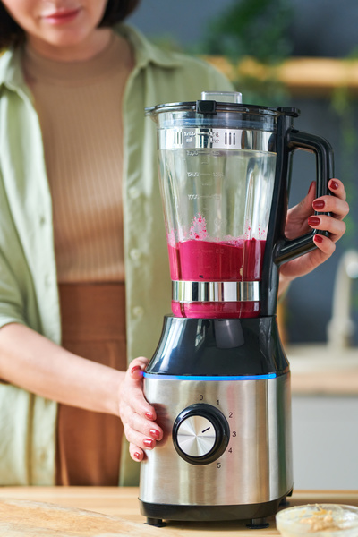 Berries and fruits are ground in a stationary blender that a smoothie maker with short hair in a light-colored outfit holds