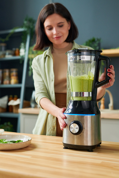 A smoothie maker with short hair and dressed in light-colored clothes is making a green spinach smoothie in a stationary blender keeping it on the table