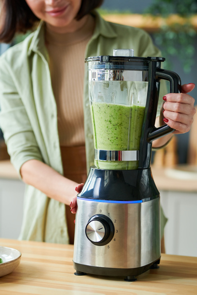 Fruits and spinach are crushed on a table made of light wood stationary blender with a black handle held by a woman making smoothie