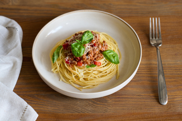Pasta with bolognese sauce and basil leaves lies in a white plate on a wooden table next to a white cloth napkin and fork