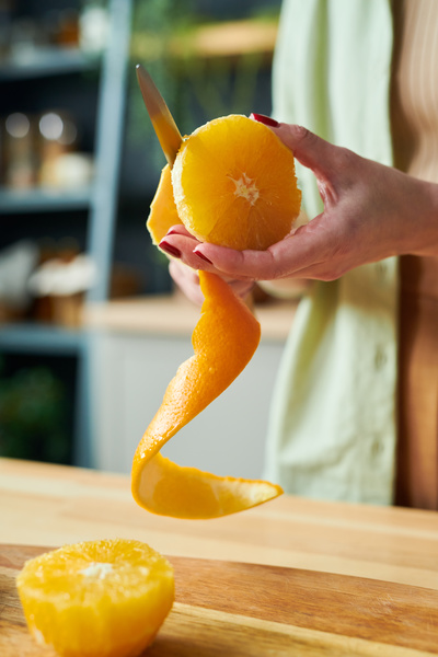 A woman cuts an orange peel with a small knife over a table with an already peeled half of an orange