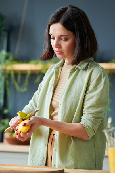 A smoothie maker with dark short-cropped hair wearing a light green shirt over a top removes mango peel with a small knife