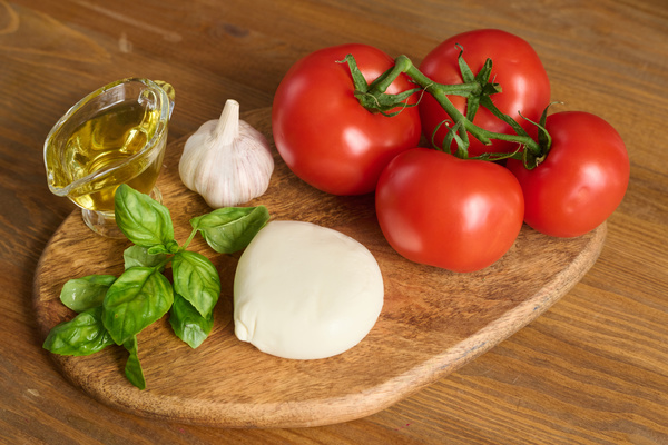 The ingredients for the salad tomatoes on a sprig a head of garlic a gravy boat of vegetable oil basil and mozzarella cheese lie on a wooden board