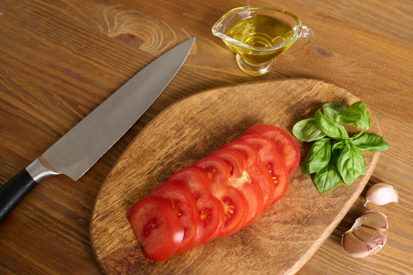 Sliced tomatoes and basil leaves lie on a cutting board next to a gravy boat butter chef's knife and half a head of garlic